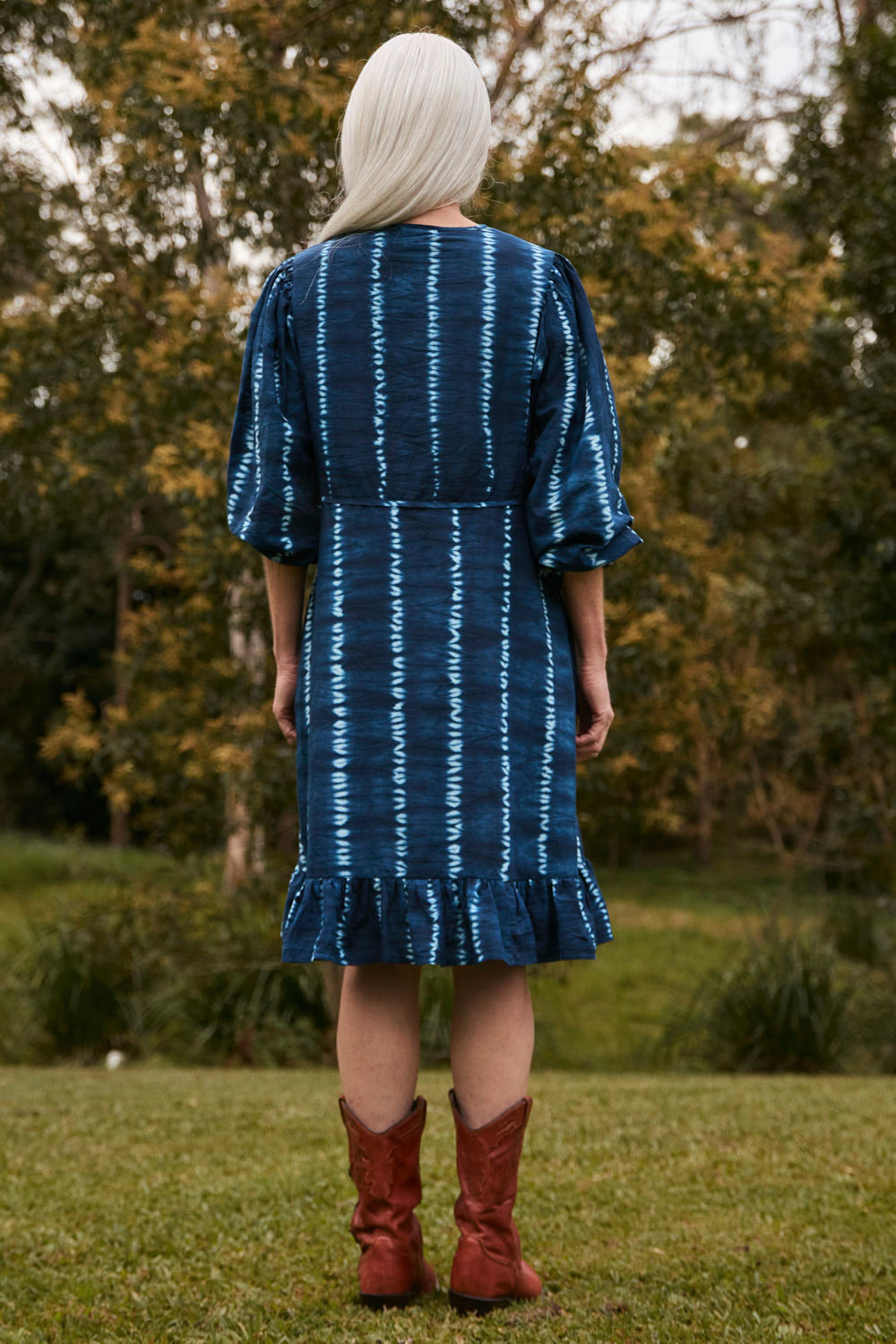 Person wearing a blue and white patterned dress with red boots standing in a grassy area with trees.