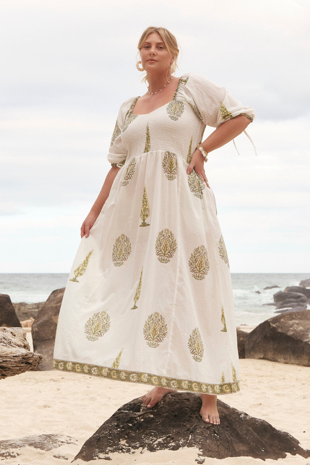 Woman in a white dress with gold patterns standing on a rocky beach.