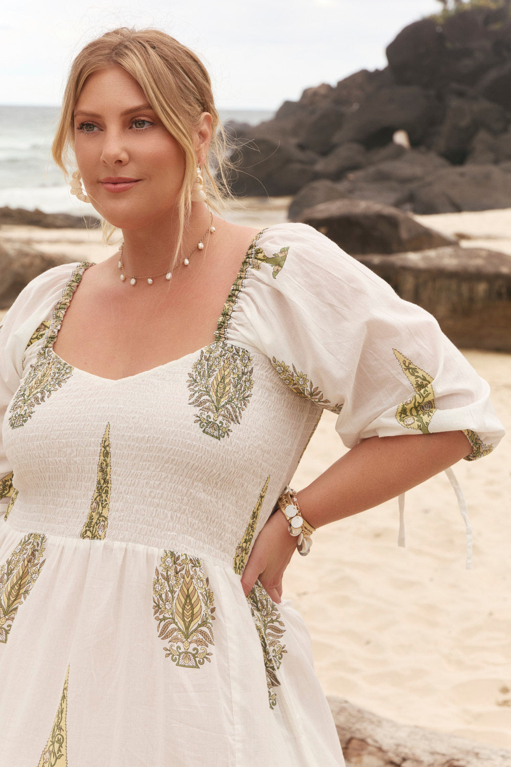 Woman in a white dress with green patterns standing on a beach.