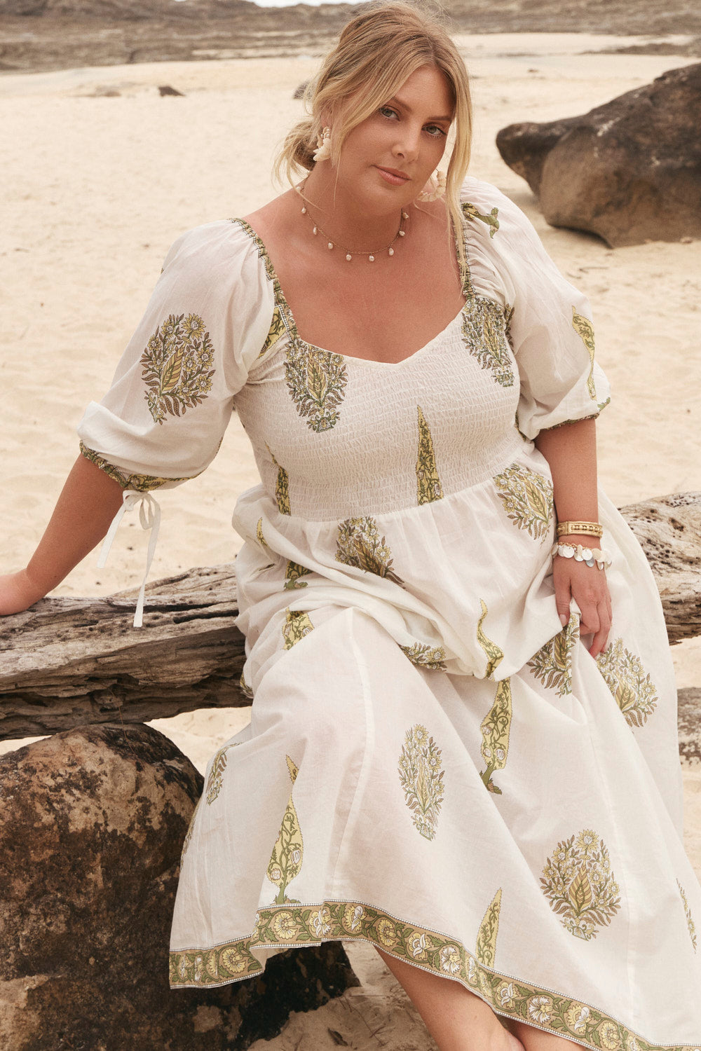 Woman in a white dress with green patterns sitting on a beach.