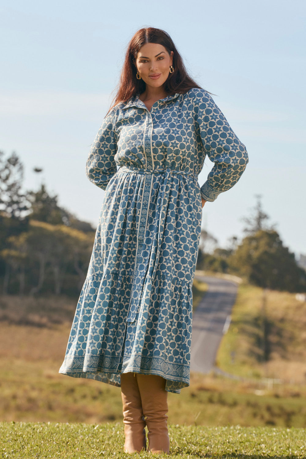 Woman wearing a blue patterned dress standing outdoors with trees and a clear sky in the background