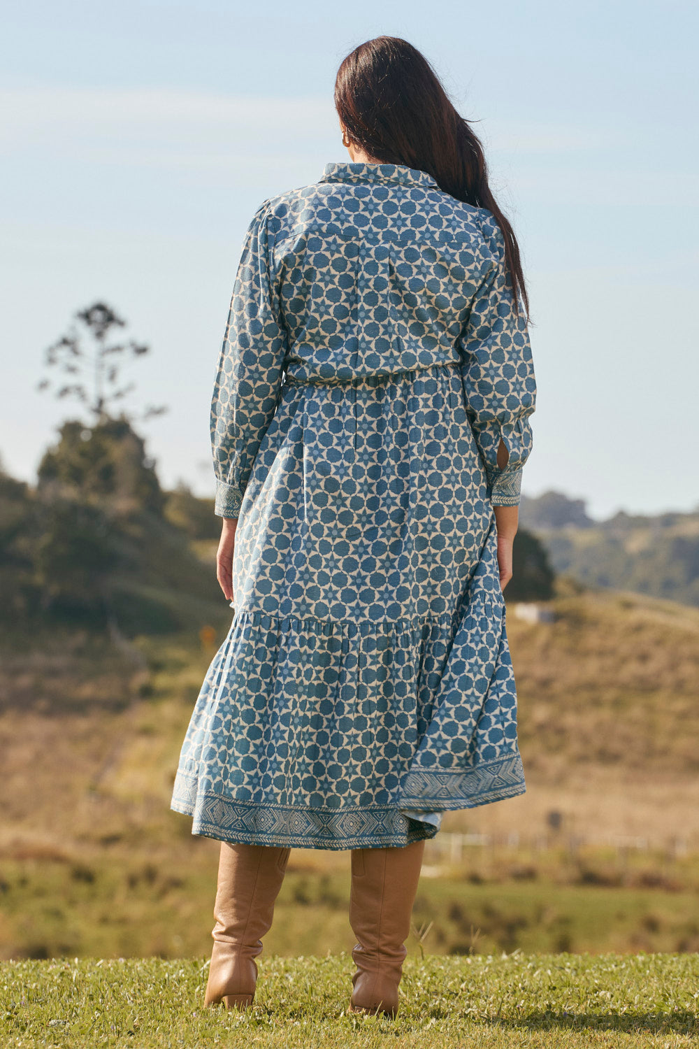 Person wearing a blue patterned dress standing in a grassy field with trees in the background