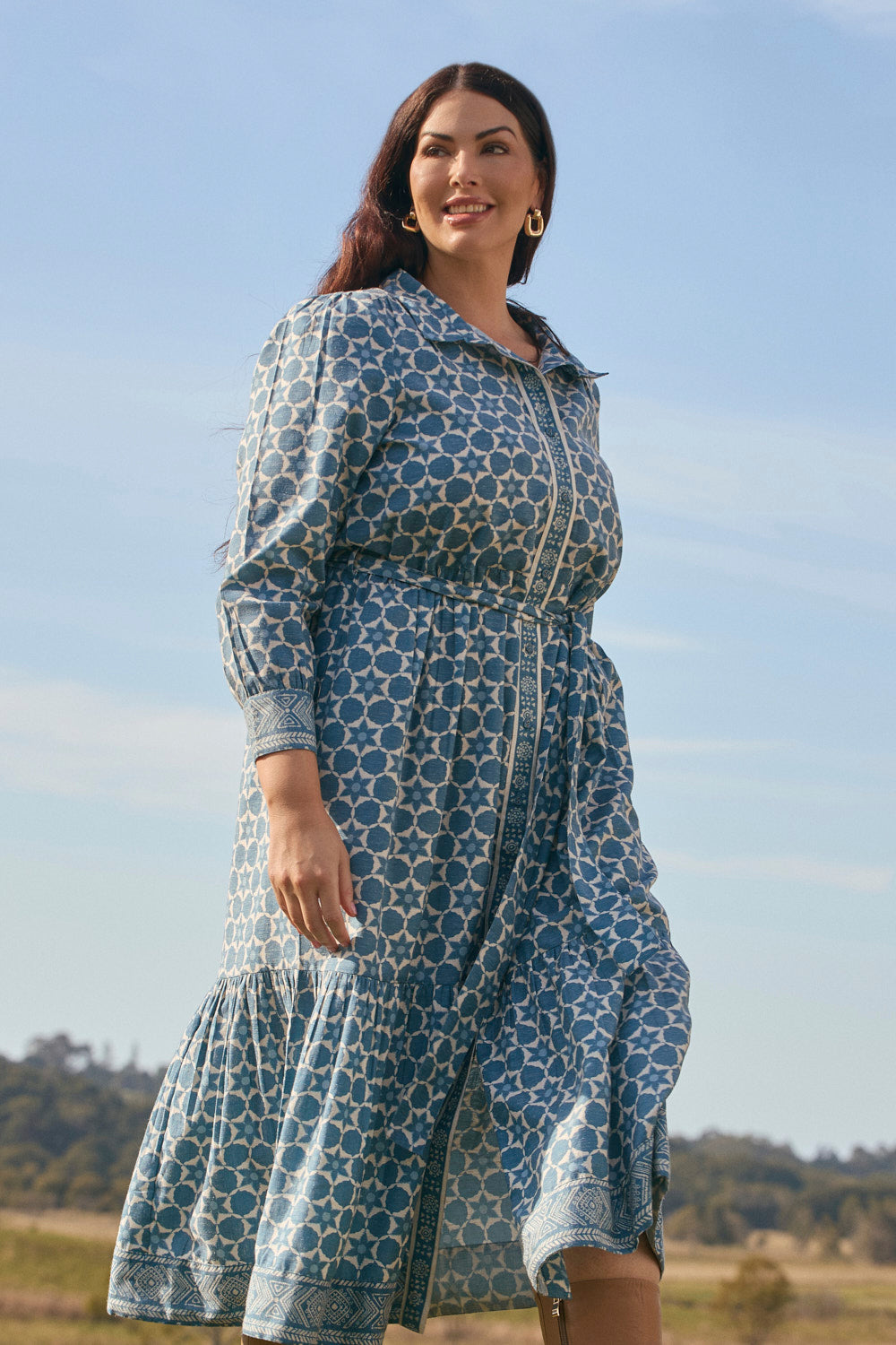 Woman wearing a blue patterned dress standing outdoors with a clear sky and greenery in the background