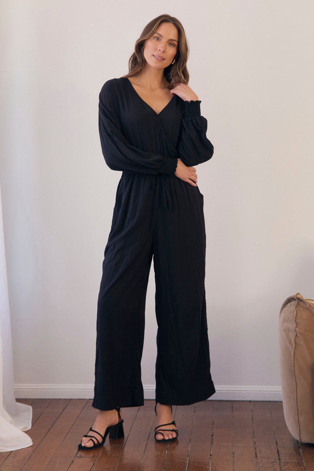 Woman wearing a black jumpsuit standing in a room with a white wall and wooden floor.
