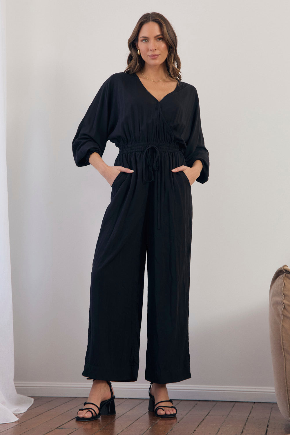 Woman wearing a black jumpsuit standing against a white wall.