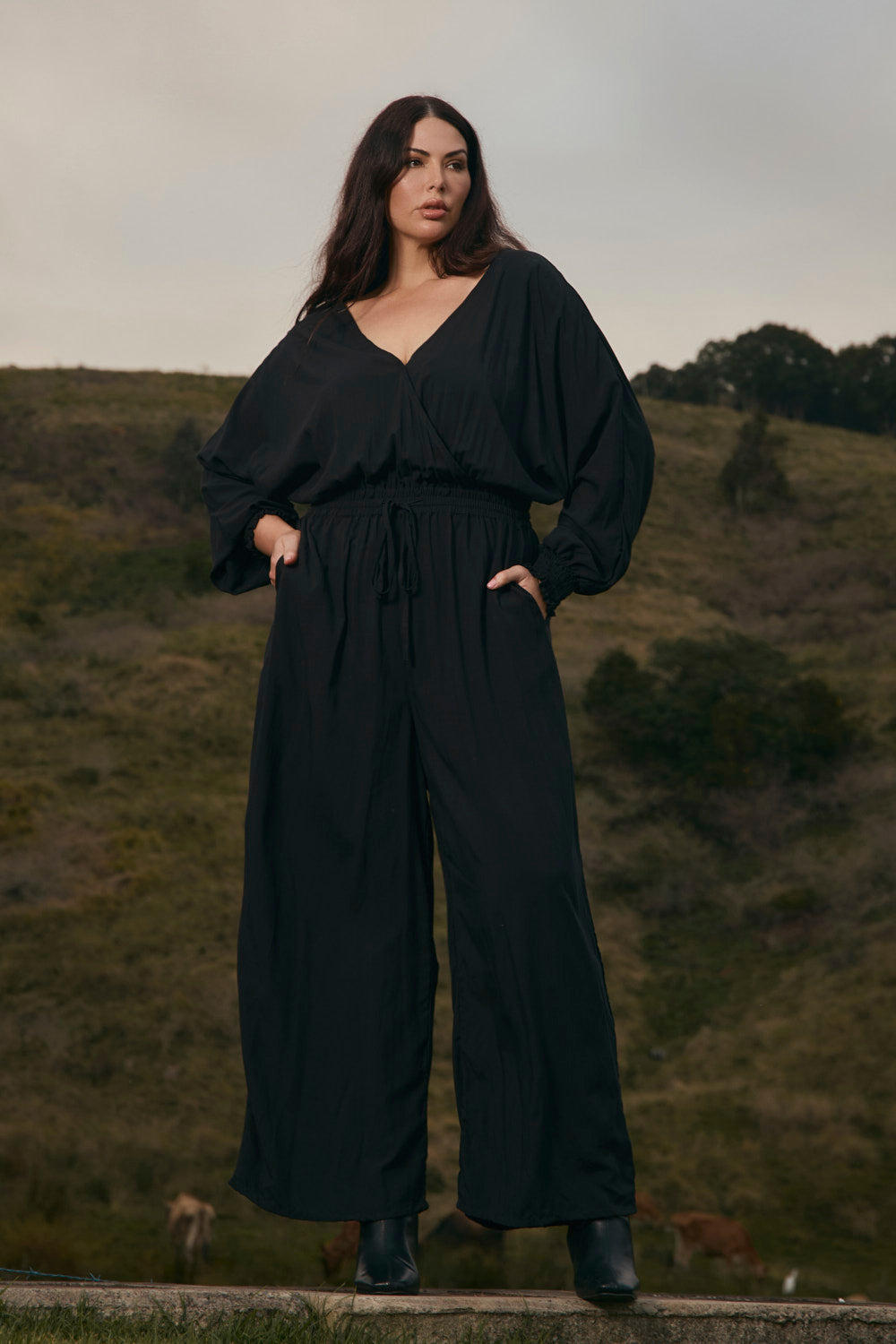 Woman wearing a black jumpsuit standing in a natural setting with greenery.