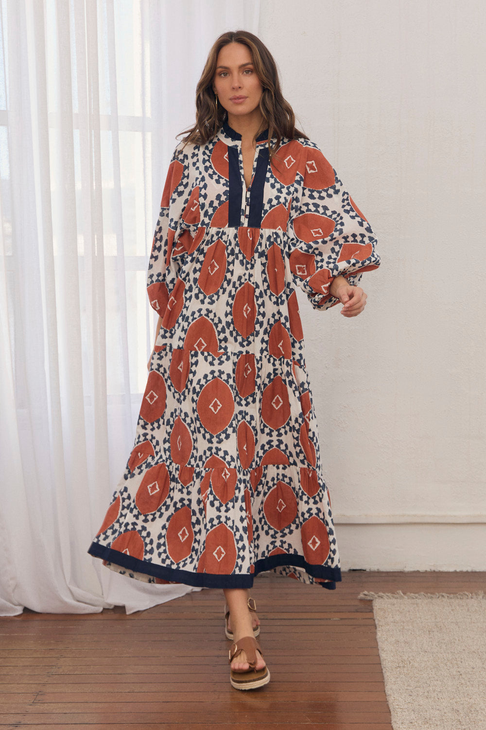 Woman wearing a long, patterned dress standing in a room with white curtains.