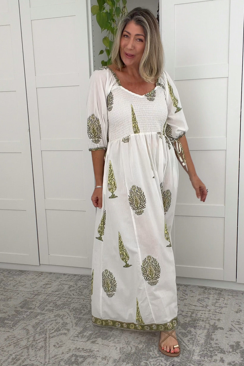 Woman wearing a white dress with green patterns in a room with white paneled walls.