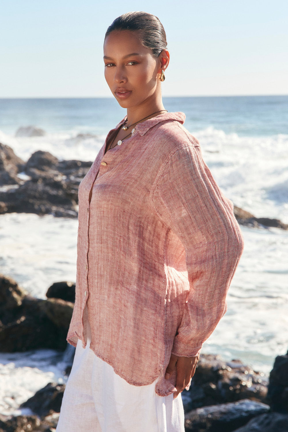 Woman standing on a rocky beach wearing a pink shirt and white pants.