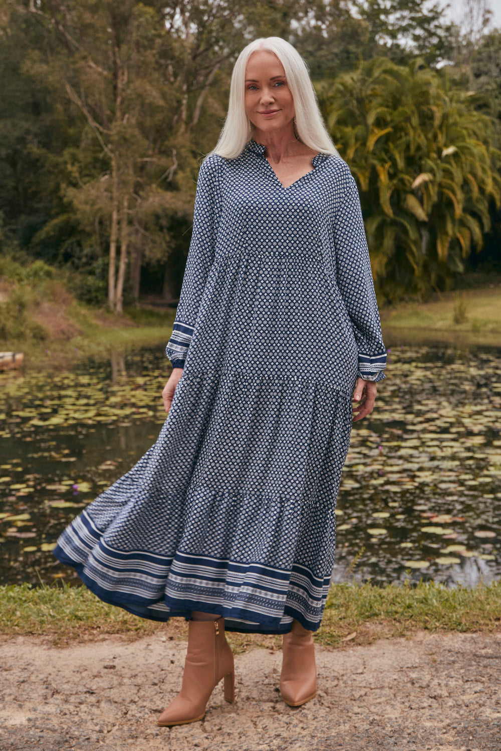 Woman in a blue patterned dress standing by a pond with greenery in the background