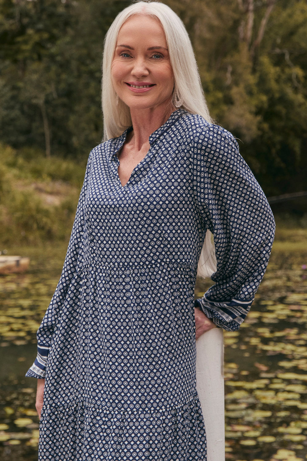 Woman wearing a blue patterned dress standing by a body of water with greenery in the background