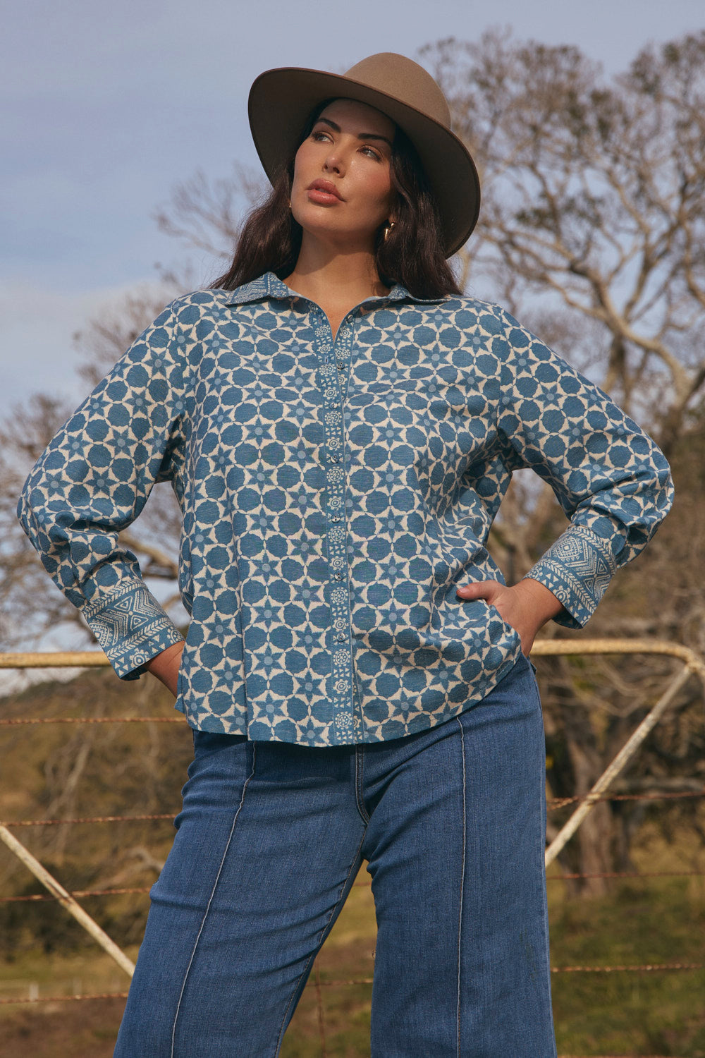 Woman wearing a patterned shirt and jeans standing outdoors with trees in the background