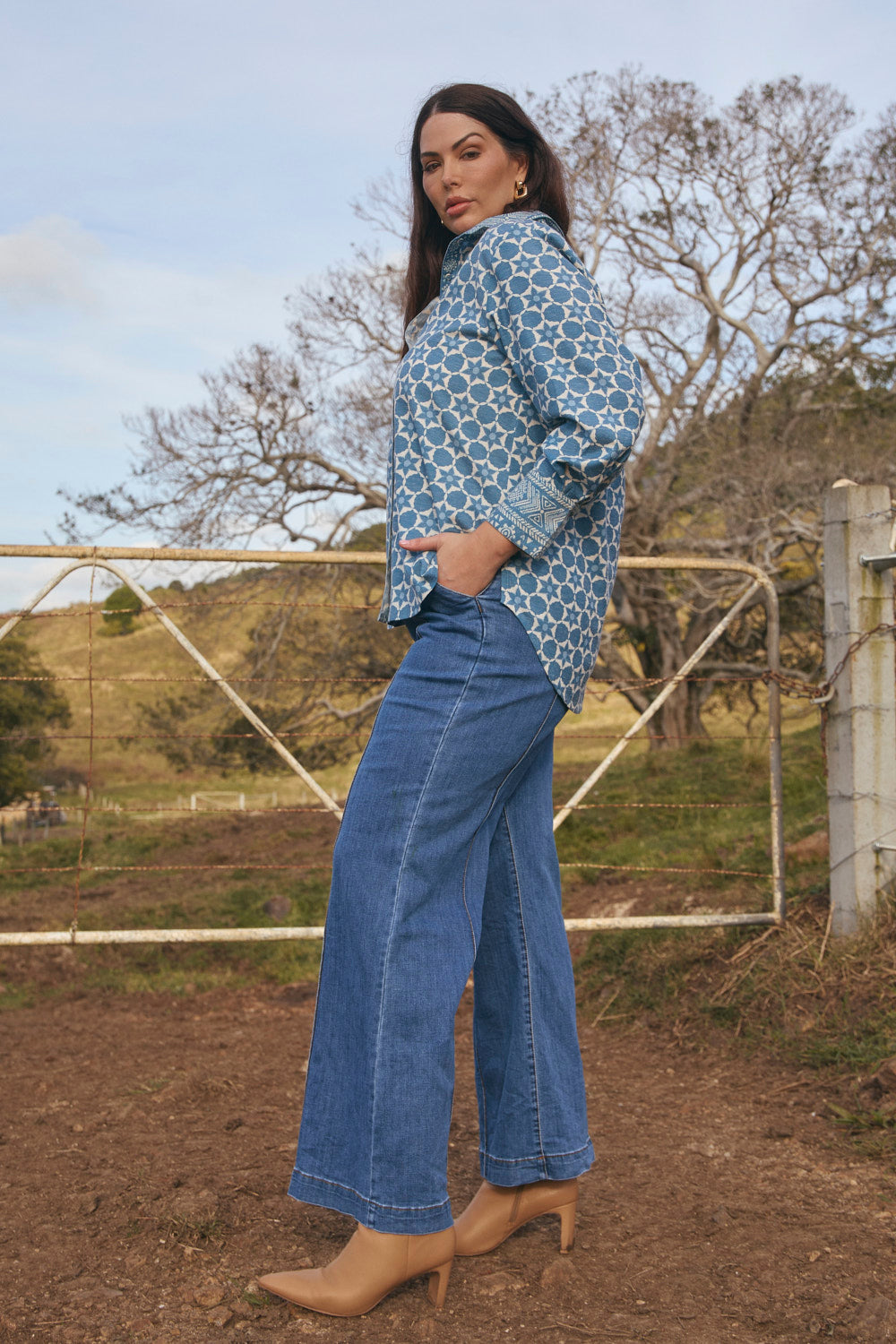 Woman wearing a patterned blouse and blue jeans standing outdoors with trees and a fence in the background.