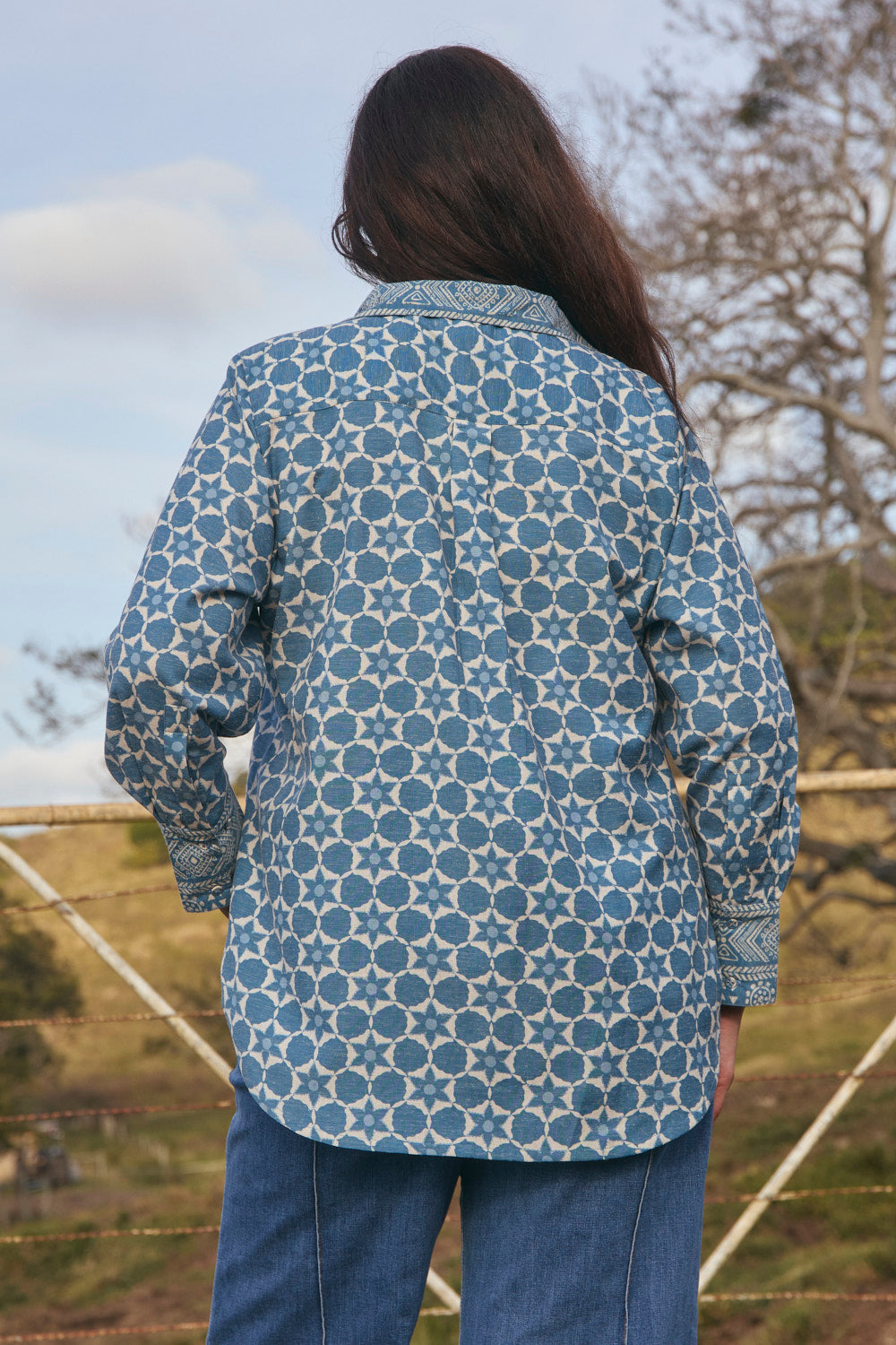 Person wearing a blue patterned shirt outdoors with trees and sky in the background