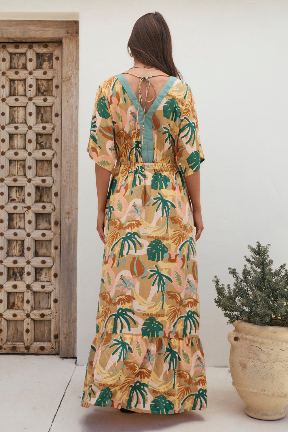 Woman wearing a tropical patterned dress standing in front of a wooden door and potted plant.