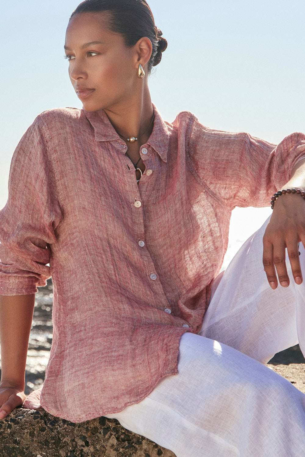 Woman wearing a pink shirt and white pants sitting on a rock by the beach.